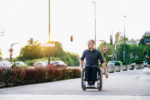 Ein Mann im Rollstuhl ist auf dem Weg zur Arbeit. Er fährt auf einem Gehweg entlang einer Straße. Im Hintergrund geht die Sonne auf.