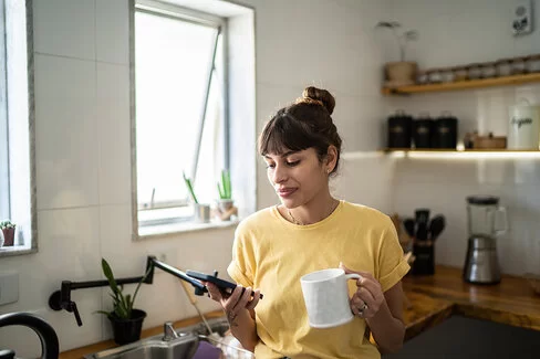 Eine junge Frau mit dunklen Haaren steht in der Küche und schaut auf ihr Smartphone, in der linken Hand hält sie einen weißen Bescher. Sie lächelt.