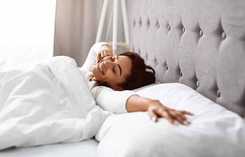 Eine junge Frau liegt in ihrem Bett in einem lichtdurchfluteten Schlafzimmer. Sie streckt nach dem Aufwachen ihre Arme aus.