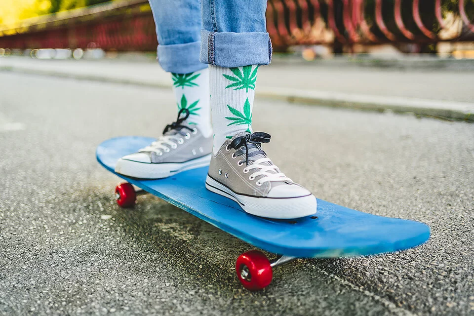 Im Bildausschnitt sind die Beine eines Menschen zu sehen. Er trägt Blue Jeans und graue Sneaker. Auf den weißen Socken sind grüne Marihuana-Pflanzen zu sehen. Er steht auf einem blauen Skateboard und fährt eine Straße entlang.