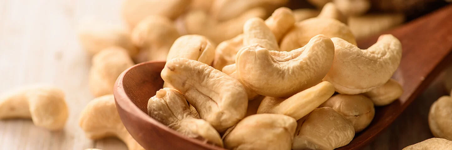 Naturbelassene Cashewkerne liegen in einem Holzlöffel und auf einem Tisch.