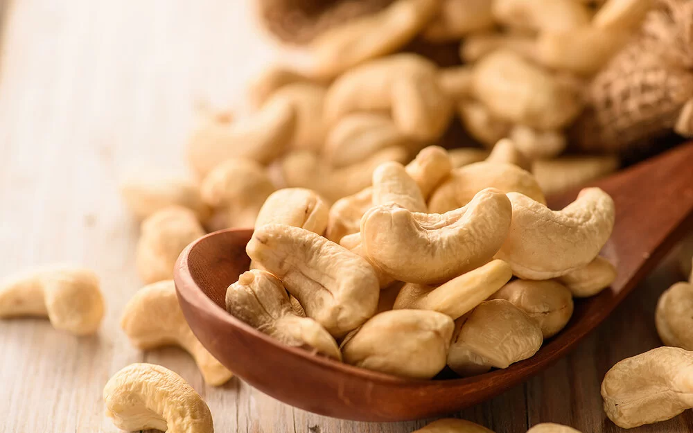 Naturbelassene Cashewkerne liegen in einem Holzlöffel und auf einem Tisch.