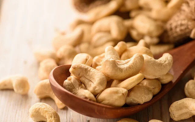 Naturbelassene Cashewkerne liegen in einem Holzlöffel und auf einem Tisch.