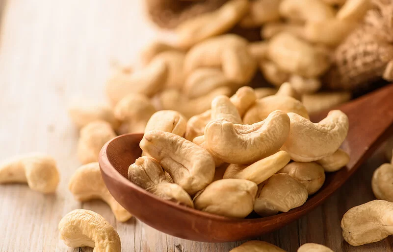 Naturbelassene Cashewkerne liegen in einem Holzlöffel und auf einem Tisch.