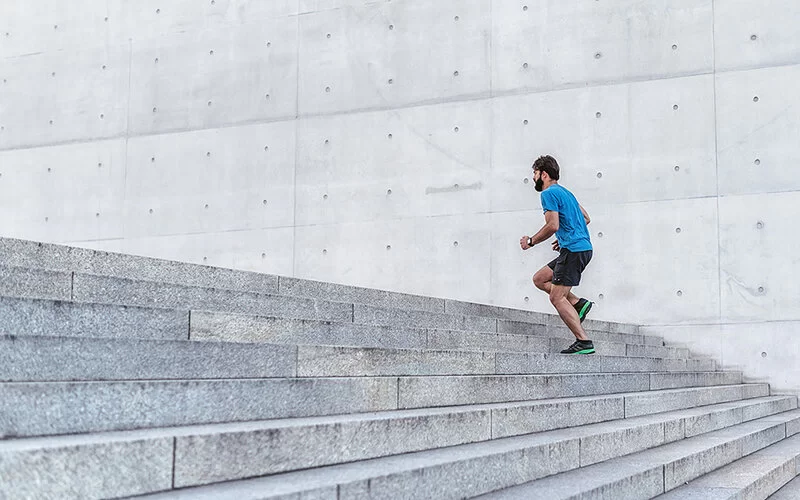 Ein junger, bärtiger Jogger läuft in kurzer Trainingskleidung eine Betontreppe im Freien hinauf.