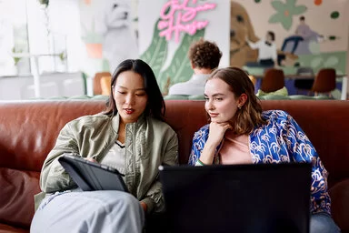 Zwei junge Frauen sitzen auf einem braunen Ledersofa und schauen auf ein Tablet, das eine der Frauen in der Hand hält. Beide Frauen haben dunkle schulterlange Haare, eine trägt eine olivgrüne dünne Jacke, die andere eine blaue Jacke mit Palmenmuster.