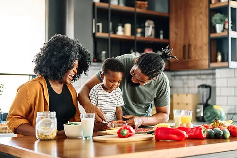 Eltern kochen mit ihrem kleinen Sohn in der Küche. Sie stehen vor einer Küchenzeile, auf der viel Gemüse liegt.