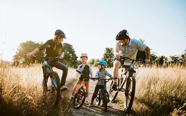 Eine Familie, bestehend aus Vater, Mutter und zwei kleinen Jungen, auf Fahrrädern, die eine Pause auf einem Feldweg einlegen. Alle tragen Helme und lächeln sich gegenseitig fröhlich an.