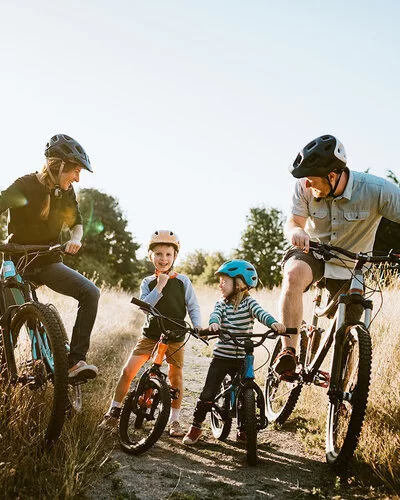 Eine Familie, bestehend aus Vater, Mutter und zwei kleinen Jungen, auf Fahrrädern, die eine Pause auf einem Feldweg einlegen. Alle tragen Helme und lächeln sich gegenseitig fröhlich an.