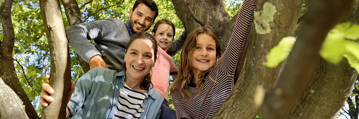 Eine Familie mit zwei Kindern ist in einen Baum geklettert und lächelt nun herunter in die Kamera. Der dunkelhaarige Vater trägt ein graues Hemd und einen Vollbart. Die Mutter trägt ein schwarz-weiß gestreiftes T-Shirt und darüber ein offenes Jeanshemd, ihre dunkelbraunen Haare trägt sie in einem Pferdeschwanz. Die beiden Töchter sind ebenfalls dunkelhaarig. Eine trägt ein rosa T-Shirt, die andere ein rosa-schwarz gestreiftes Langarm-Shirt, eine dunkelrote Shorts und eine rosa Strumpfhose. 