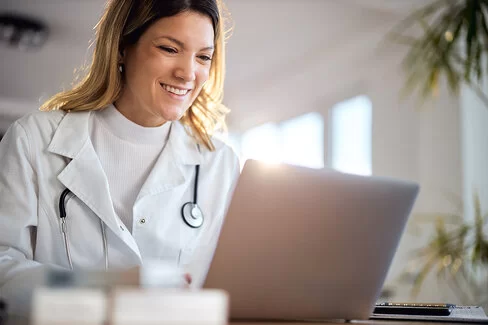 Eine Ärztin in weißer Kleidung und langen blonden Haaren sitzt vor einem Laptop in der Praxis und führt lächelnd eine Videokonferenz.