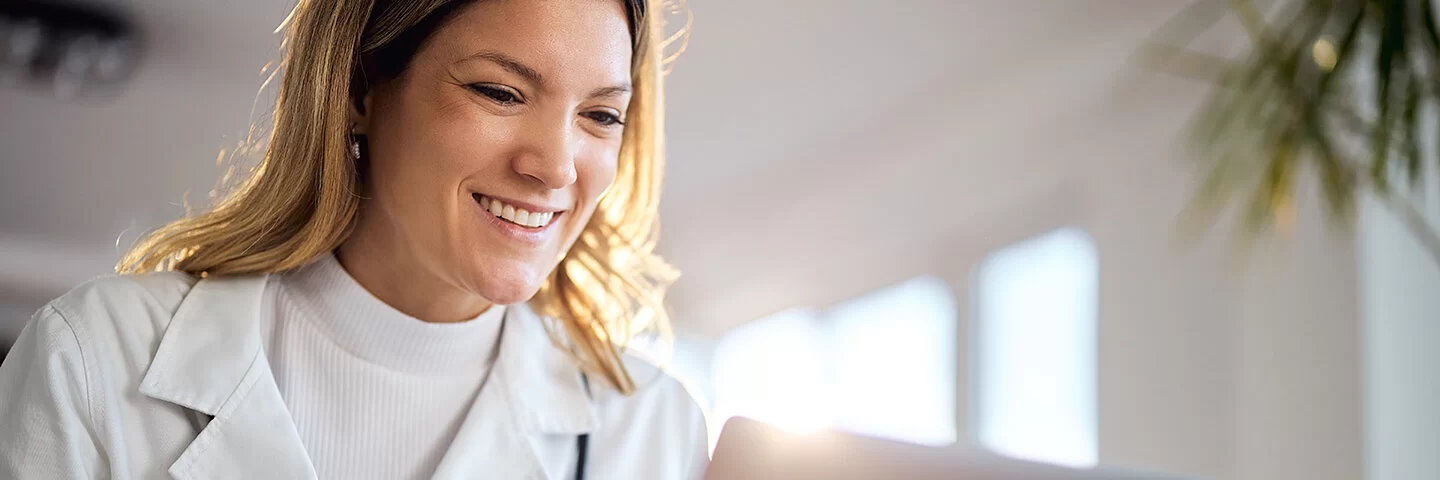 Eine Ärztin in weißer Kleidung und langen blonden Haaren sitzt vor einem Laptop in der Praxis und führt lächelnd eine Videokonferenz.
