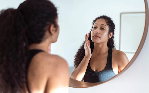 Eine Frau mit langen lockigen Haaren in schwarzem Trägertop steht vor einem runden Spiegel und ist im Begriff, ihr Gesicht mit den Fingern zu berühren.