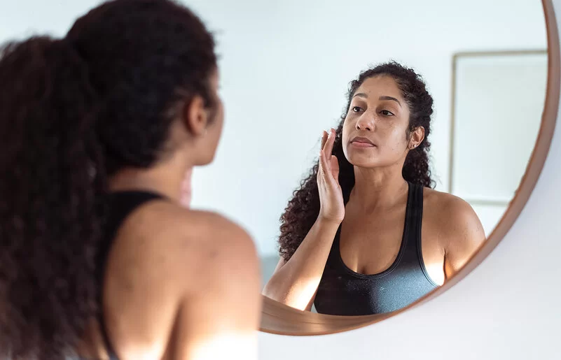 Eine Frau mit langen lockigen Haaren in schwarzem Trägertop steht vor einem runden Spiegel und ist im Begriff, ihr Gesicht mit den Fingern zu berühren.