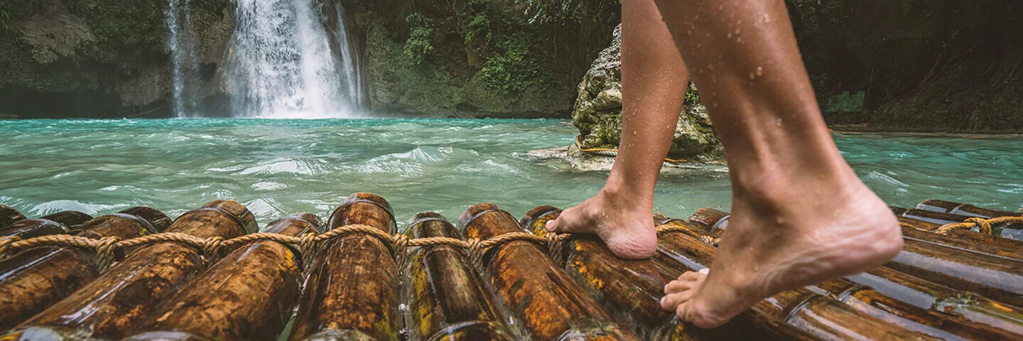 Nahaufnahme von nackten Frauenunterschenkeln, die auf einem nassen Bambusfloß stehen, das auf einen Wasserfall zusteuert.