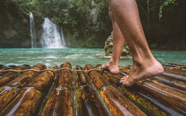 Nahaufnahme von nackten Frauenunterschenkeln, die auf einem nassen Bambusfloß stehen, das auf einen Wasserfall zusteuert.