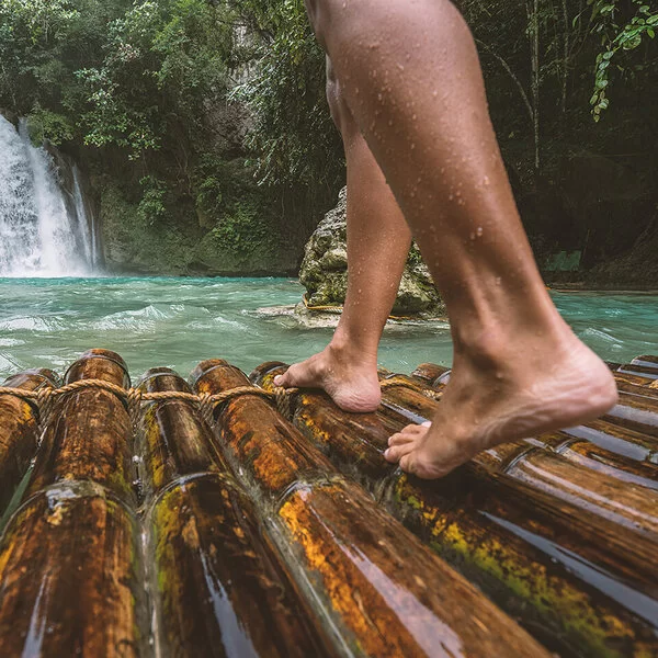 Nahaufnahme von nackten Frauenunterschenkeln, die auf einem nassen Bambusfloß stehen, das auf einen Wasserfall zusteuert.