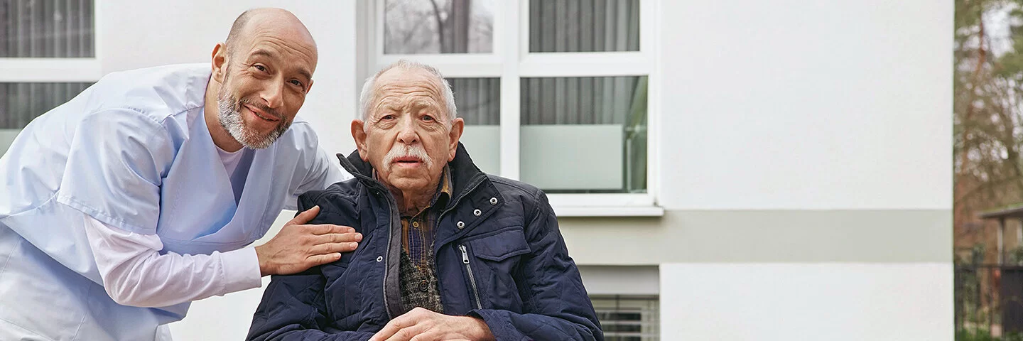 Ein älterer Mann mit dunkelblauer Jacke sitzt in einem Rollstuhl auf einer Terrasse, auf seinen Beinen liegt eine Decke. Eine Pflegefachkraft in hellblauer Kleidung beugt sich zum Mann herunter, eine Hand ruht auf der Schulter des Mannes. Beide lächeln in die Kamera.