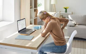 Eine Frau mit einem braunen Pullover und einer Jeanshose bekleidet, sitzt auf einem Stuhl vor einem Schreibstisch. Sie hält sich mit einer Hand den Nacken und mit der anderen Hand den Rücken.