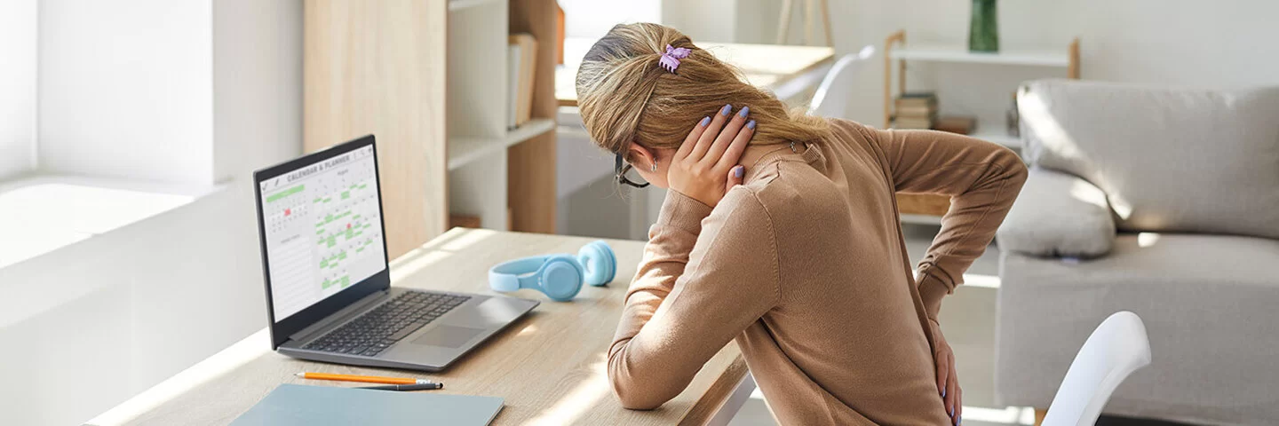 Eine Frau mit einem braunen Pullover und einer Jeanshose bekleidet, sitzt auf einem Stuhl vor einem Schreibstisch. Sie hält sich mit einer Hand den Nacken und mit der anderen Hand den Rücken.