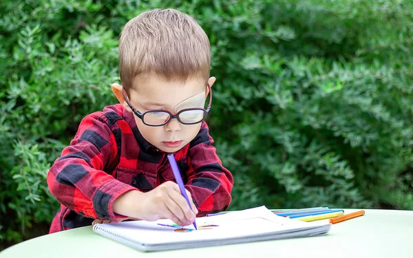Ein Junge mit Amblyopie hat sein linkes Auge mit einem Okkluder abgeklept, er tr�ägt dazu eine Brille und sitzt malend im Garten an einem Tisch.