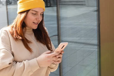 Eine junge Frau mit langem dunklen Haar, gelber Wollmütze und beigefarbenen Pullover steht vor einem Gebäude in der Stadt und schaut auf ihr Smartphone.