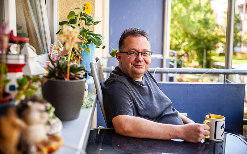 Jörg Lohse sitzt auf dem Balkon und lächelt. Mit seinen Händen umfasst er eine Tasse, die auf einem runden Tisch steht. Einige Blumentöpfe stehen auf der Fensterbank.