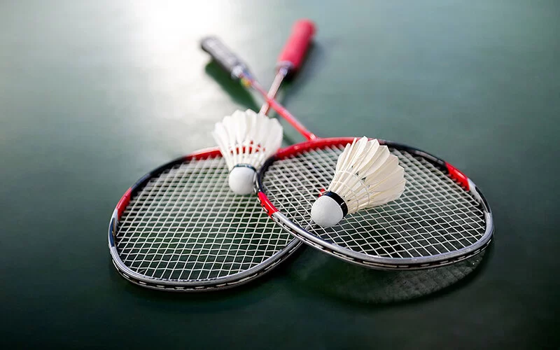 Zwei Federbälle und zwei Badminton-Schläger liegen auf einem dunkelgrünen Linoleumboden.