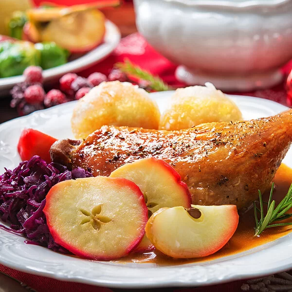 Auf einem festlichen gedeckten Tisch steht ein weißer Teller. Darauf sind eine gebratene Entenkeule, Rotkohl, Kartoffelknödel, Apfelscheiben und ein kleiner, frischer Zweig Rosmarin angerichtet.