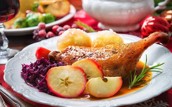 Auf einem festlichen gedeckten Tisch steht ein weißer Teller. Darauf sind eine gebratene Entenkeule, Rotkohl, Kartoffelknödel, Apfelscheiben und ein kleiner, frischer Zweig Rosmarin angerichtet.