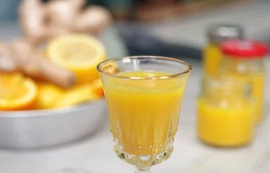 Ingwer-Shot nach einem Rezept von Felicitas Then, serviert in einem Kristallglas. Rechts im Hintergrund weitere Shots in Schraubgläsern. Links im Hintergrund eine silberne Schale voller Ingwerknollen, Zitronen und Orangen.