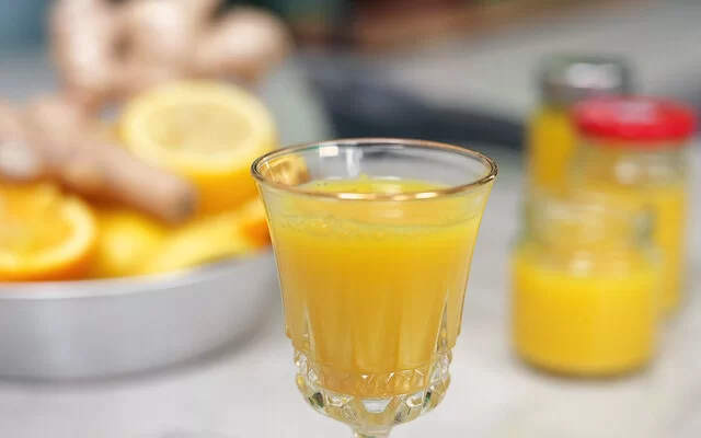 Ingwer-Shot nach einem Rezept von Felicitas Then, serviert in einem Kristallglas. Rechts im Hintergrund weitere Shots in Schraubgläsern. Links im Hintergrund eine silberne Schale voller Ingwerknollen, Zitronen und Orangen.