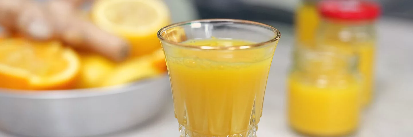 Ingwer-Shot nach einem Rezept von Felicitas Then, serviert in einem Kristallglas. Rechts im Hintergrund weitere Shots in Schraubgläsern. Links im Hintergrund eine silberne Schale voller Ingwerknollen, Zitronen und Orangen.