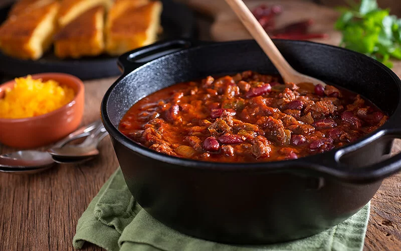 Auf einem Tisch steht ein schwarzer gusseiserner Topf mit Chili sin Carne. Dazu werden geriebener Käse und Brot gereicht.