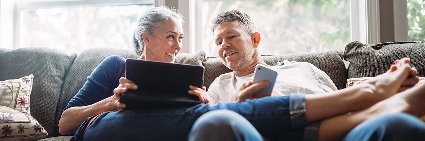 Ein Pärchen in seinen 50ern lümmelt gemeinsam auf der Couch – sie hält ein Tablet, er ein Smartphone in Händen.