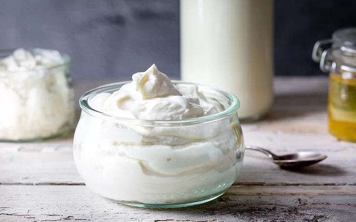 Auf einem Holztisch steht eine Glasschüssel mit Quark. Im Hintergrund befinden sich ein weiteres Glas mit Frischkäse, eine Milchflasche und ein Honigglas.