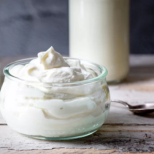 Auf einem Holztisch steht eine Glasschüssel mit Quark. Im Hintergrund befinden sich ein weiteres Glas mit Frischkäse, eine Milchflasche und ein Honigglas.