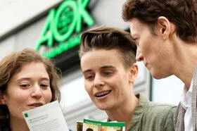 Eine Frau präsentiert zwei jungen Frauen Prospekte vor einer AOK-Filiale. 