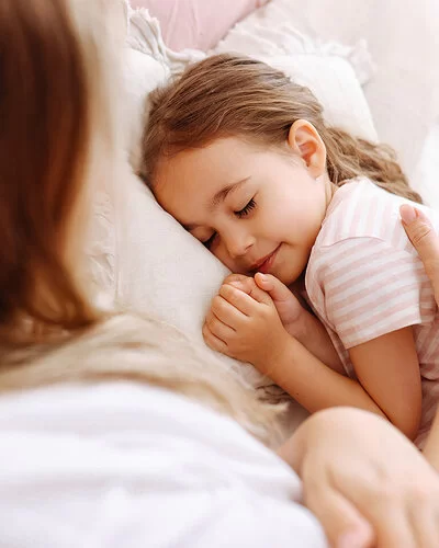 Ein ungefähr fünfjähriges Mädchen liegt im Bett und schläft. Ihre Mutter streicht ihr mit dem rechten Arm fürsorglich über den Rücken.