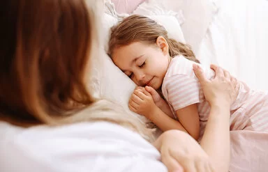 Ein ungefähr fünfjähriges Mädchen liegt im Bett und schläft. Ihre Mutter streicht ihr mit dem rechten Arm fürsorglich über den Rücken.