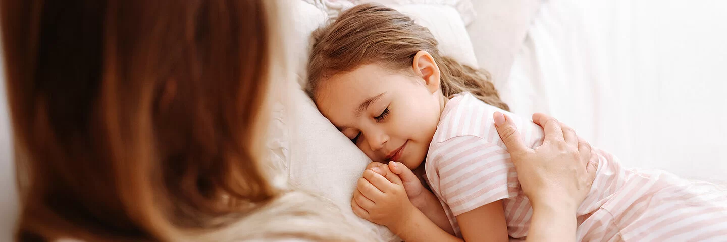 Ein ungefähr fünfjähriges Mädchen liegt im Bett und schläft. Ihre Mutter streicht ihr mit dem rechten Arm fürsorglich über den Rücken.