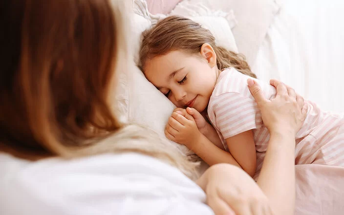 Ein ungefähr fünfjähriges Mädchen liegt im Bett und schläft. Ihre Mutter streicht ihr mit dem rechten Arm fürsorglich über den Rücken.