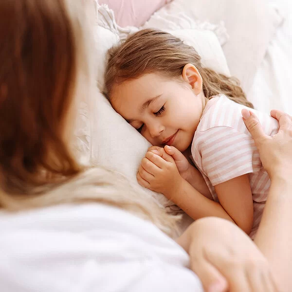 Ein ungefähr fünfjähriges Mädchen liegt im Bett und schläft. Ihre Mutter streicht ihr mit dem rechten Arm fürsorglich über den Rücken.