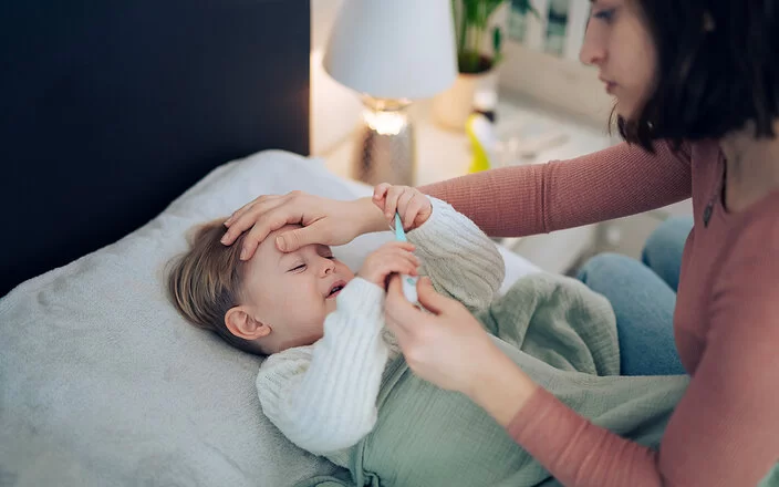 Eine Mutter nimmt die Temperatur ihres kleinen Kindes, das im Bett liegt und krank wirkt. Sie legt ihre Hand auf seine Stirn.