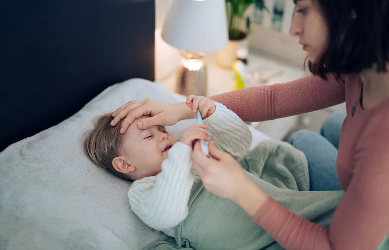 Eine Mutter nimmt die Temperatur ihres kleinen Kindes, das im Bett liegt und krank wirkt. Sie legt ihre Hand auf seine Stirn.