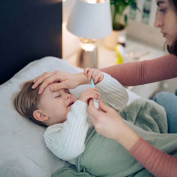 Eine Mutter nimmt die Temperatur ihres kleinen Kindes, das im Bett liegt und krank wirkt. Sie legt ihre Hand auf seine Stirn.