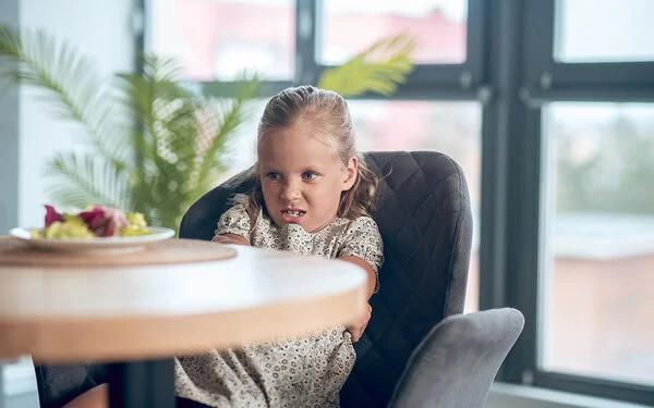 Ein kleines Mädchen in geblümtem Sommerkleid sitzt mit verschränkten Armen auf einem grauen Sessel und schaut angeekelt auf einen Teller mit Gemüse, der auf einem Tisch vor ihr steht.