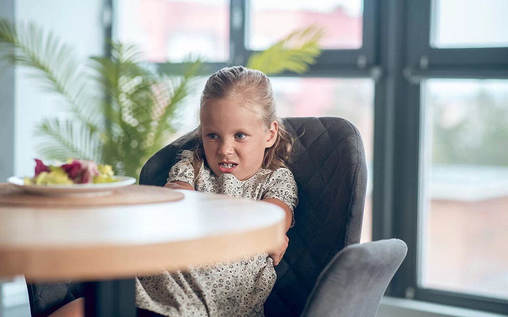 Ein kleines Mädchen in geblümtem Sommerkleid sitzt mit verschränkten Armen auf einem grauen Sessel und schaut angeekelt auf einen Teller mit Gemüse, der auf einem Tisch vor ihr steht.
