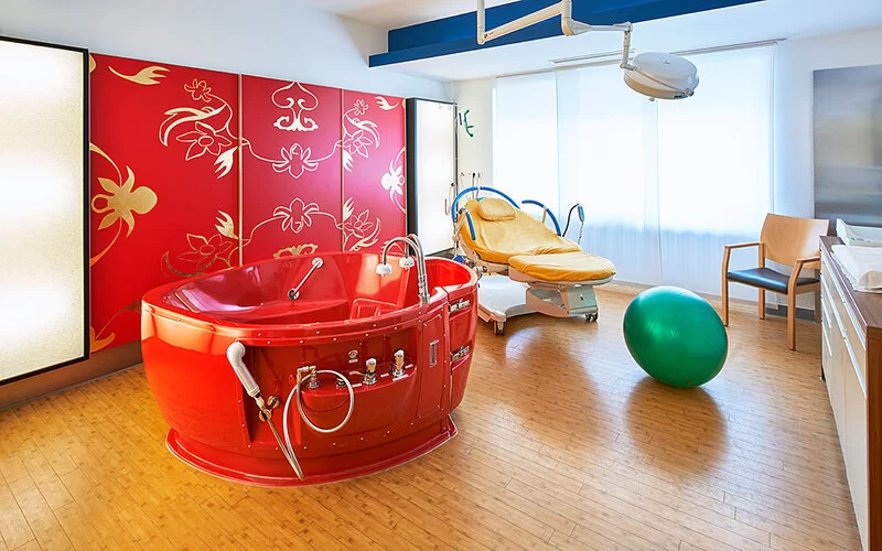 Eine rote Gebärbadewanne, auch Entbindungswanne genannt, für Wassergeburten im Kreißsaal einer Entbindungsklink, im Hintergrund steht eine Entbindungsliege. Auf dem Boden liegt ein grüner Gymnastikball.