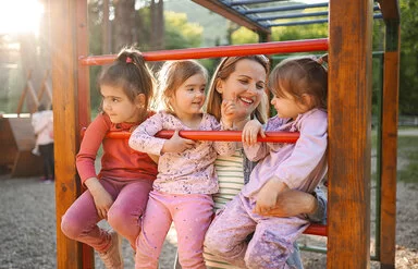 Drei Kinder sitzen lächelnd auf Klettergerüst, von einer Frau gehalten